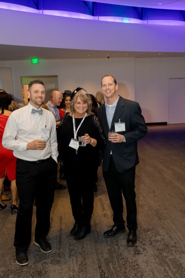 Three people standing together at an event. Man in bow tie, woman, and man in blazer smiling, holding drinks.