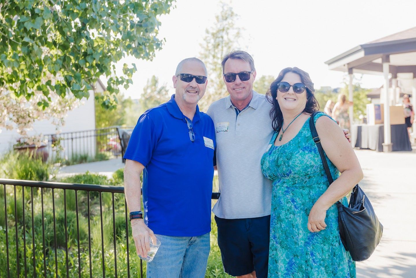 Three people pose for a photo outdoors. Man in blue shirt, man in gray shirt, woman in blue dress, all wearing sunglasses.