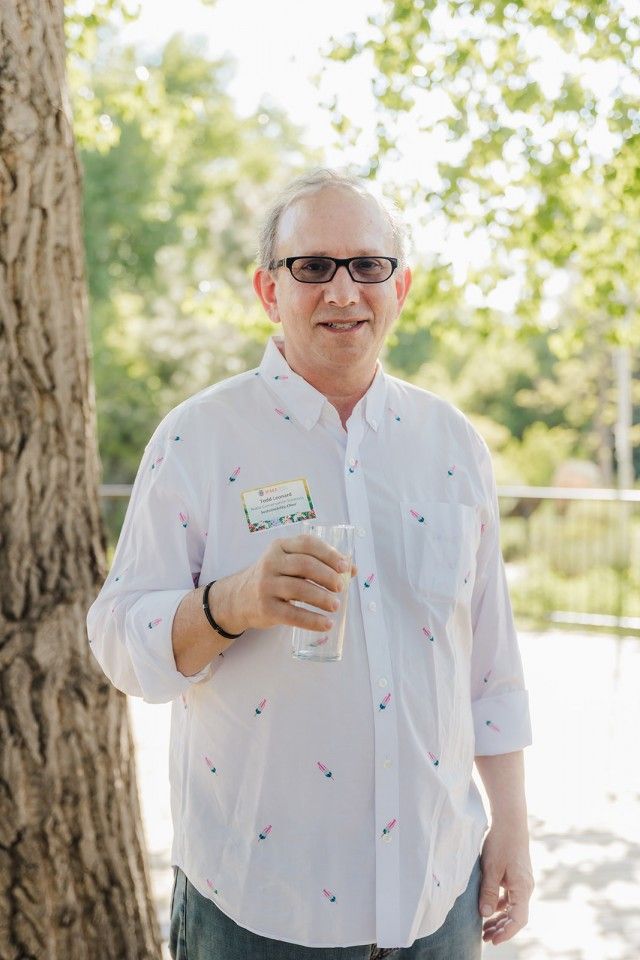 Man in glasses and patterned shirt holding a glass of water, standing outdoors near a tree.