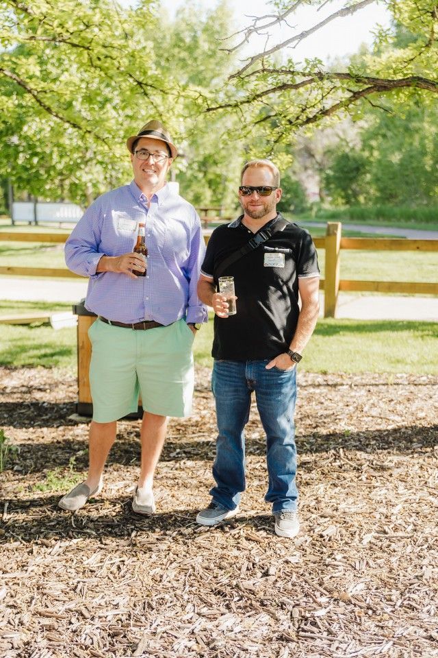 Two men outdoors holding drinks, one in blue shirt and shorts, the other in black shirt and jeans, sunny day.