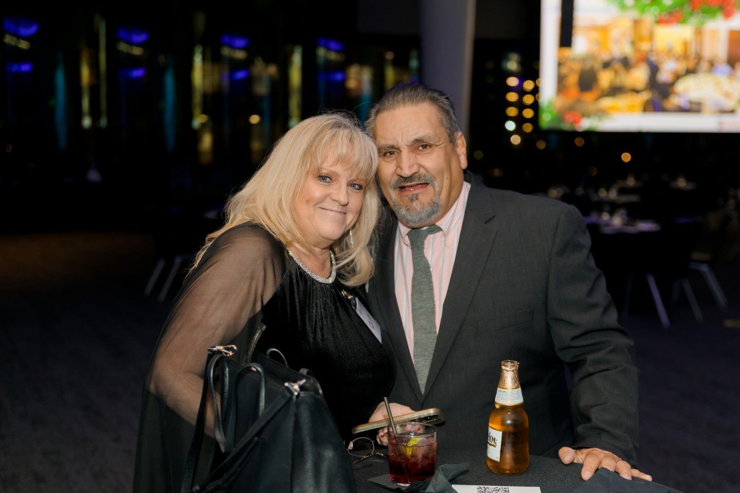 Couple smiles together at an event. Man in suit, woman in black dress, drinks and a beer on a table.