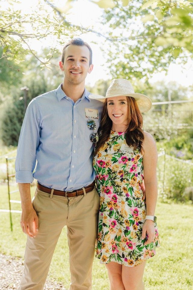 Man and woman smiling, standing outdoors. Man in blue shirt, khakis. Woman in floral dress, straw hat.