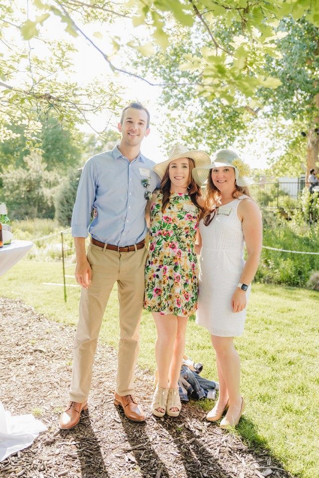 Three people smiling outdoors: man in blue shirt, woman in floral dress, woman in white dress, all wearing hats.