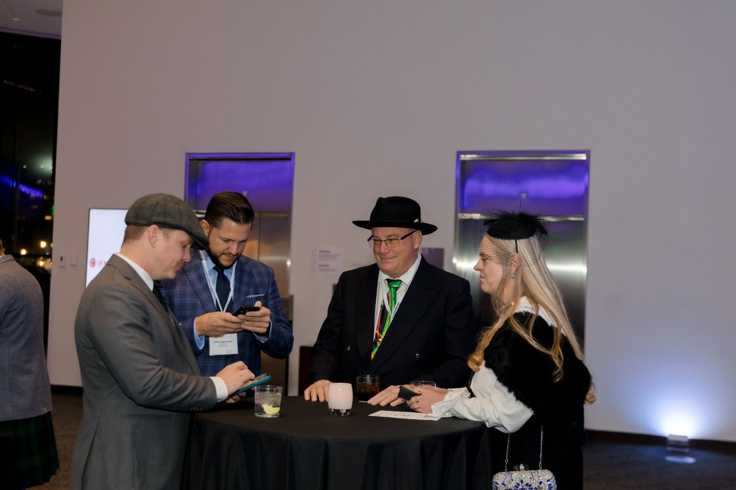 Four people converse around a small black table in a modern setting. Two men look at a phone.