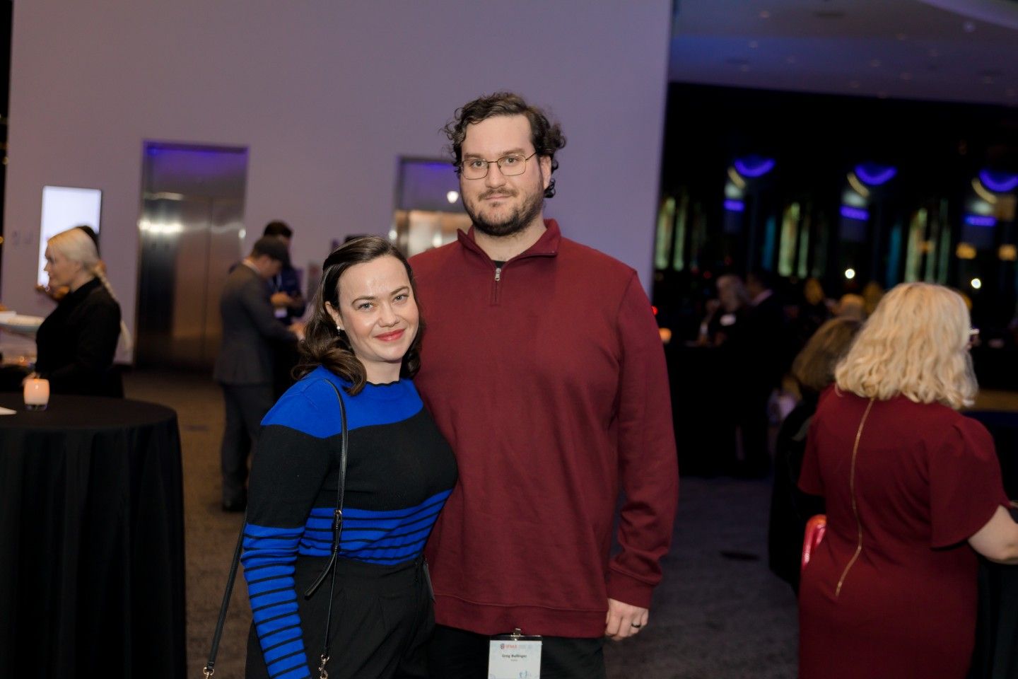 Woman in blue-striped top and man in red sweater pose together at an event. Dark background, other guests visible.