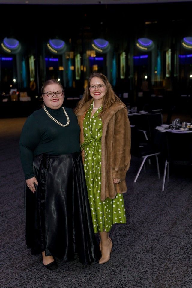 Two women at an event; one in a green dress and fur coat, the other in a black skirt and green sweater.