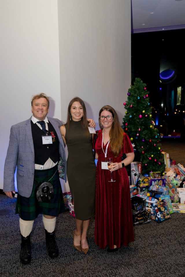 Three people stand near a Christmas tree with gifts. Man in kilt and blazer, woman in green dress, and woman in red dress.