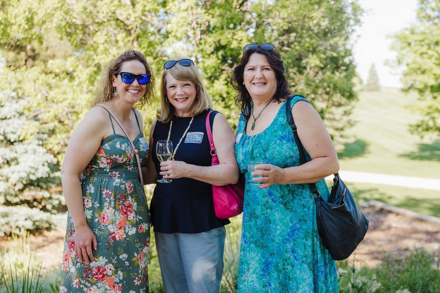 Three women smiling, standing outdoors. One in a floral dress, others in casual tops with sunglasses.