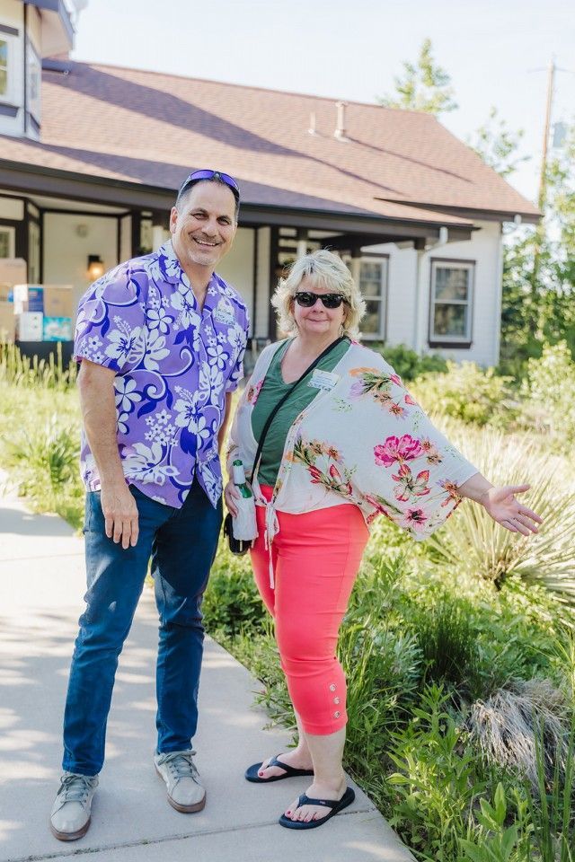 Man in floral shirt and woman in pink pants pose outdoors near a white house.