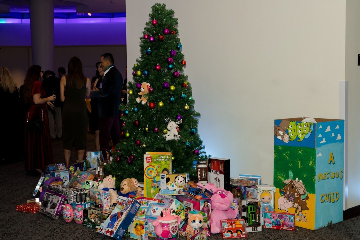Christmas tree surrounded by wrapped presents; people in the background, donation box.