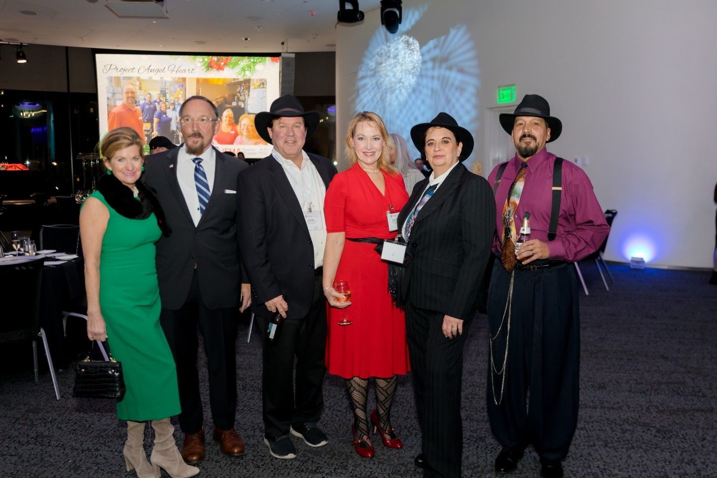 Group of people posing at an event. Some wear hats, one woman in red dress. Setting is an event hall.