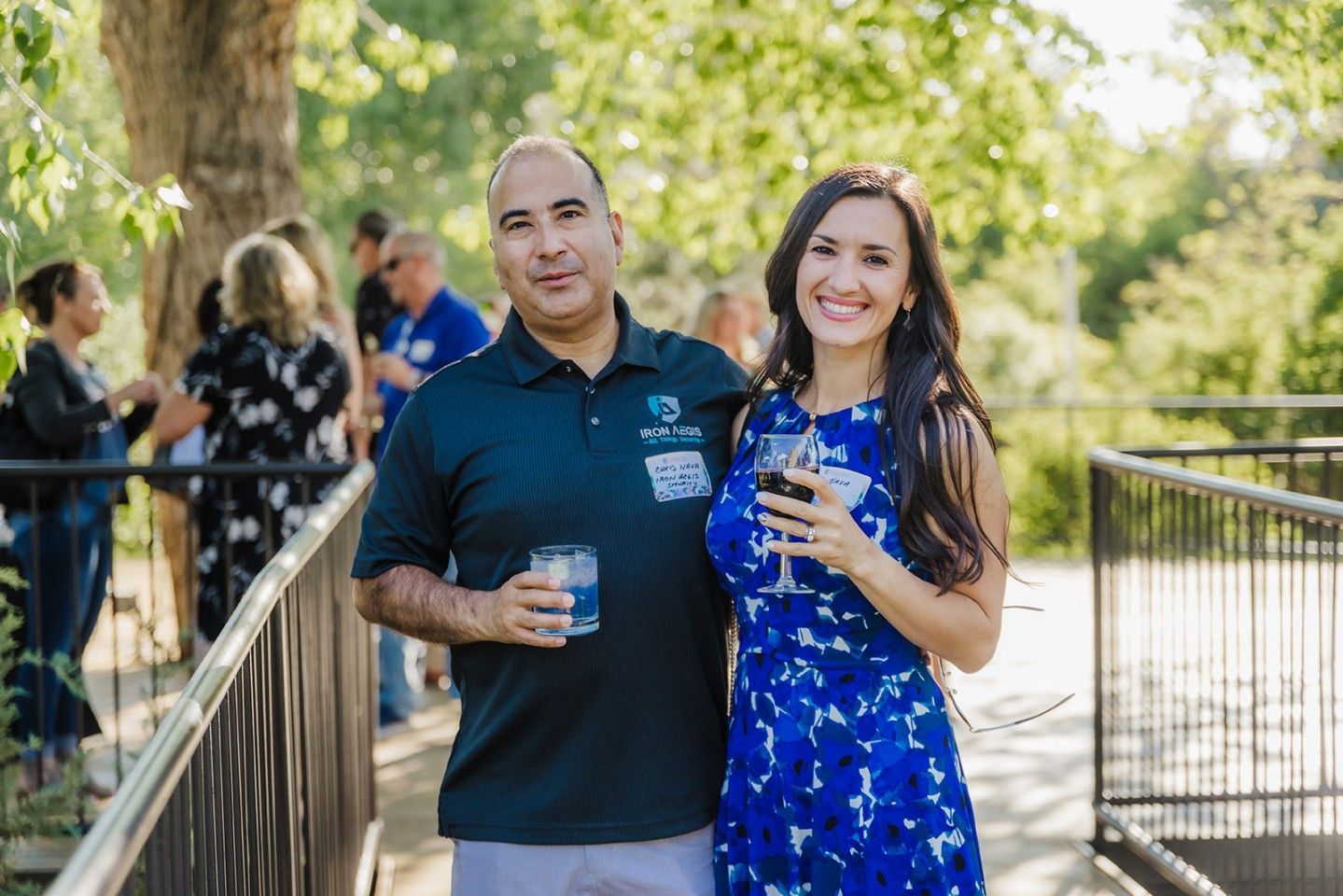 Couple smiling, holding drinks, standing on a bridge, people in background. Outdoor setting with greenery.