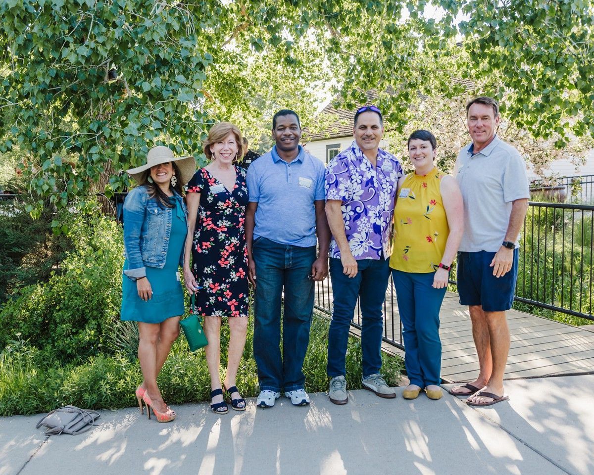 Six people standing outdoors near greenery. They are smiling and appear to be posing for a photo on a sunny day.