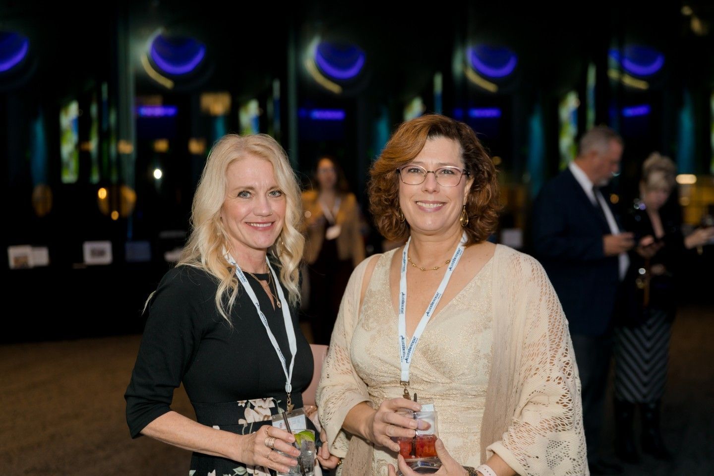 Two women smiling at an event, one in a floral dress, the other in a cream-colored dress, holding drinks.