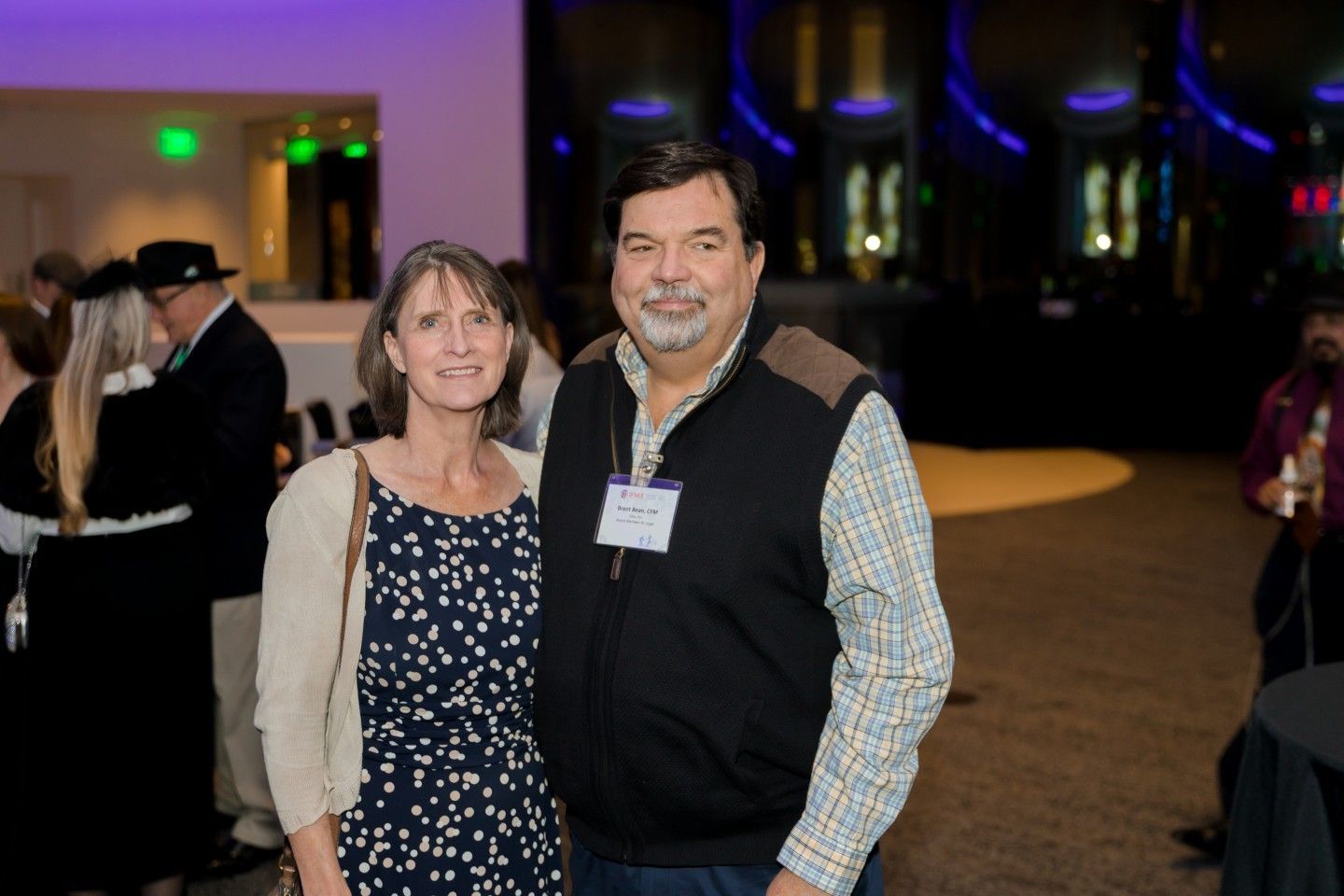 Woman and man in an event setting. The woman wears a patterned dress and sweater. The man has a name tag and vest.