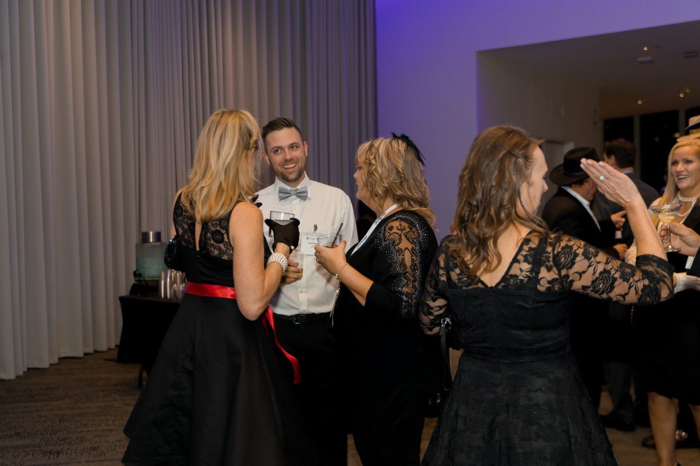 People in formal attire at an event, socializing. Some are in black dresses with lace sleeves; a man wears a bow tie.
