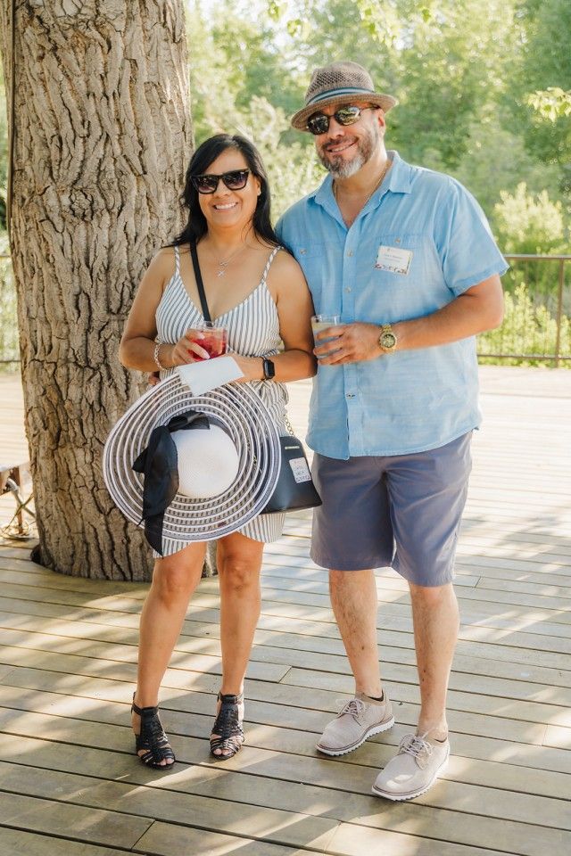 Couple smiling, posing outside. Woman in striped dress holds hat, man in blue shirt, shorts, both wear sunglasses.
