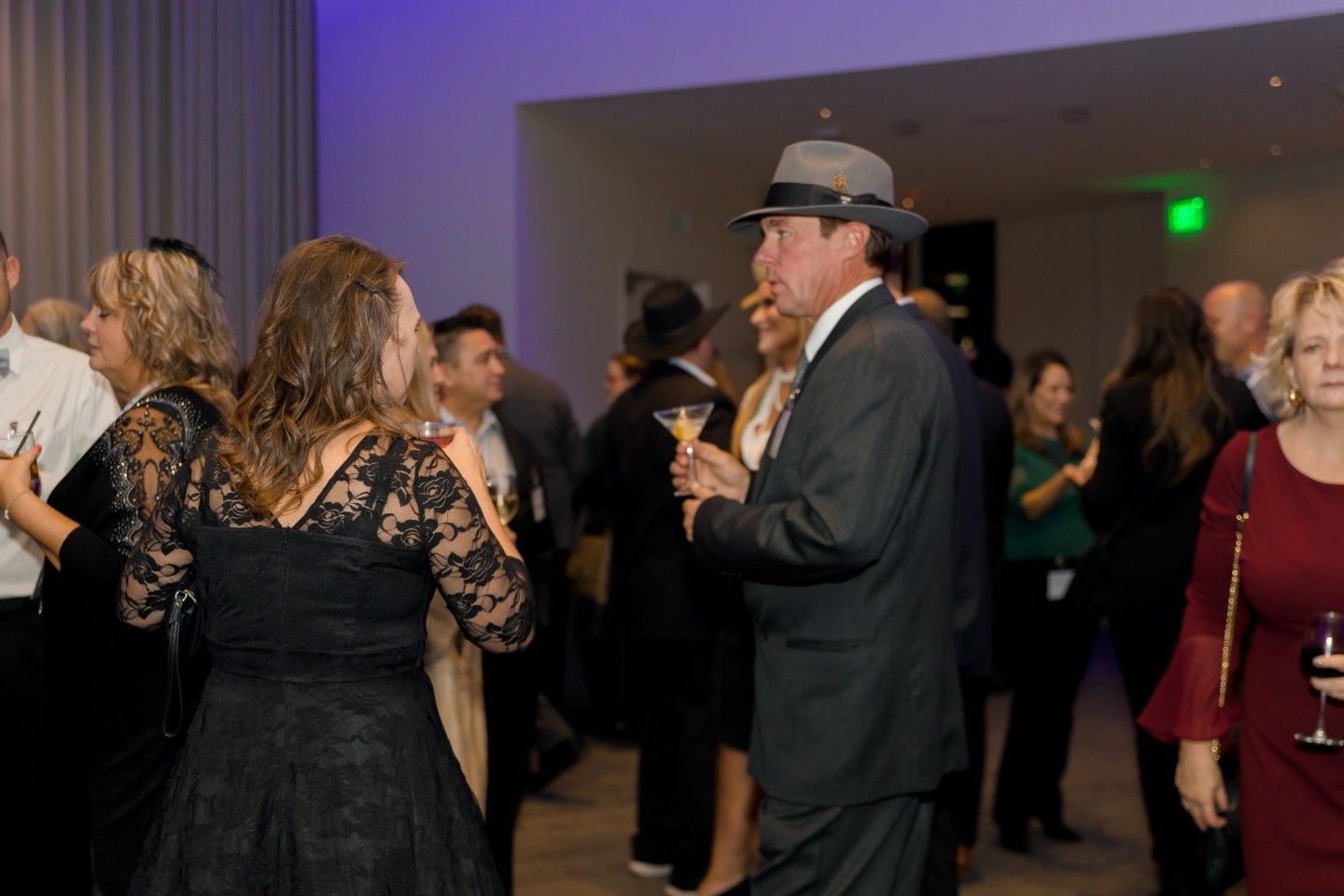 People in formal attire at an event, some wearing hats and holding drinks, indoors with soft lighting.