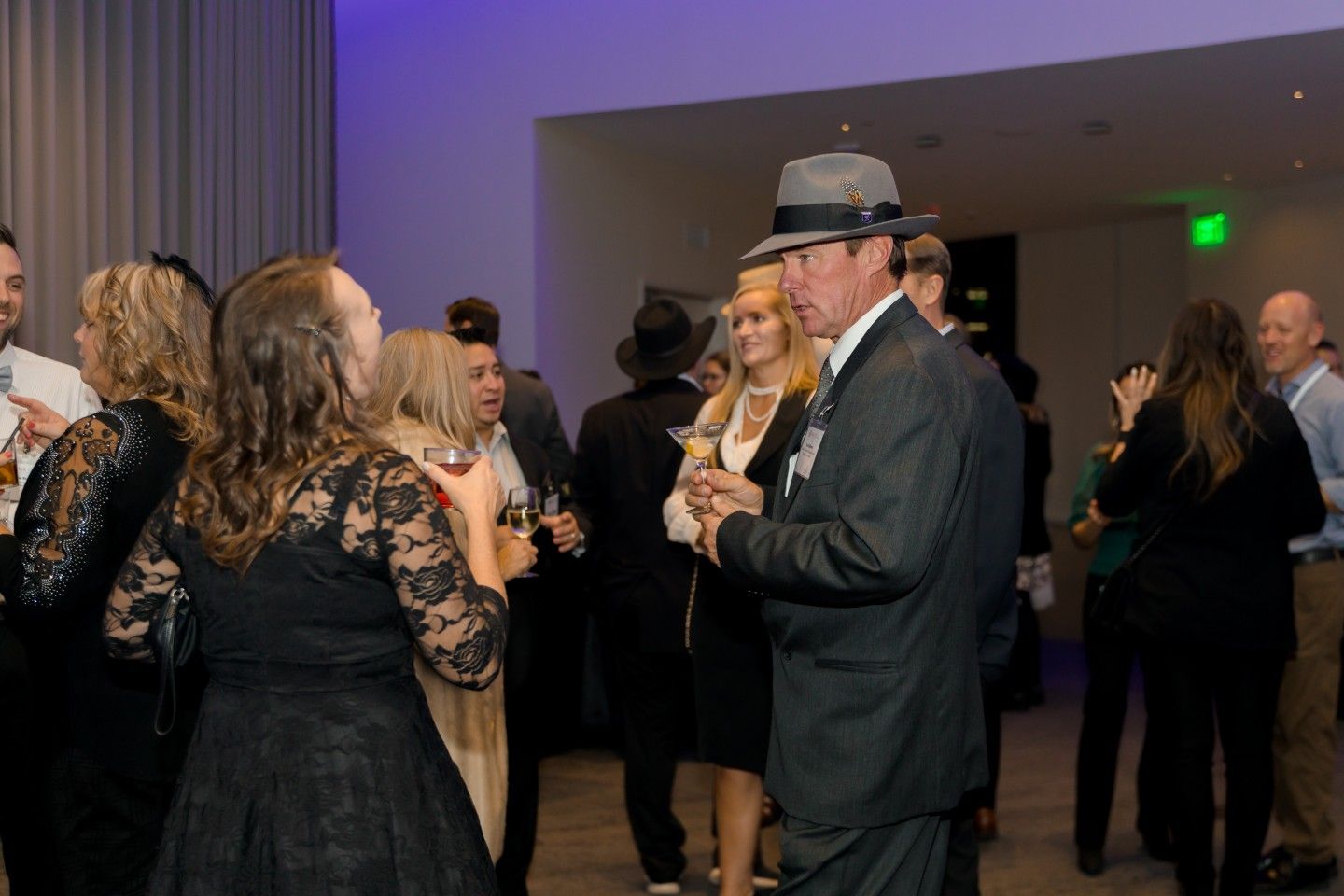 People in formal attire at an event, some are talking. Interior setting with neutral walls and lighting.