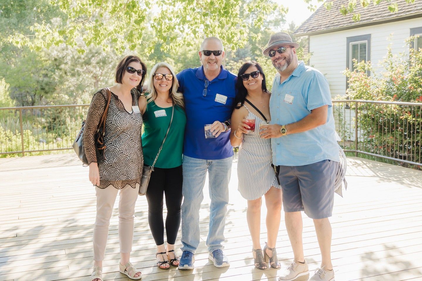 Five people smiling outdoors. Sunlight, trees, and a white building in background.