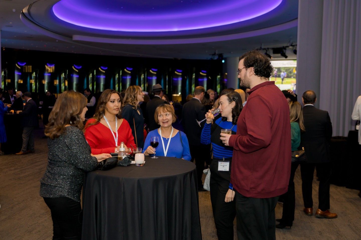 People at an event, gathered around tables, socializing, drinking, in a room with a purple ceiling.