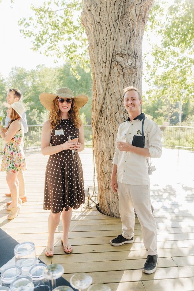 Woman in polka dots and man in chef uniform stand on a wooden deck by a tree.
