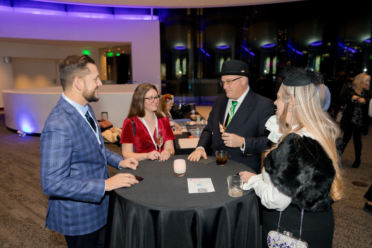 Four people at a round black table in a modern setting, socializing. One man wears a green suit and hat.