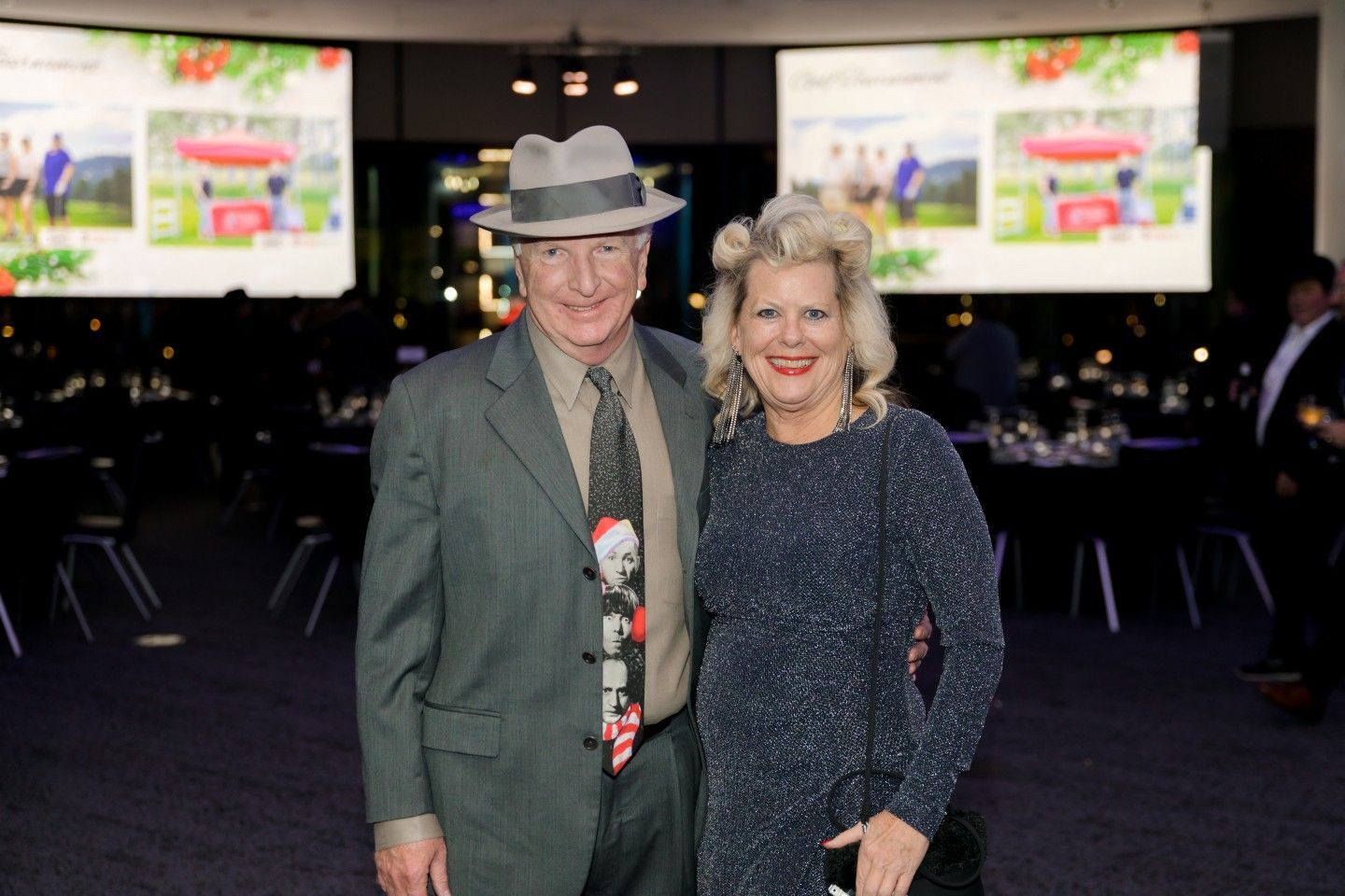 Man in hat and woman in sparkly dress pose together at a gala, with screens in the background.