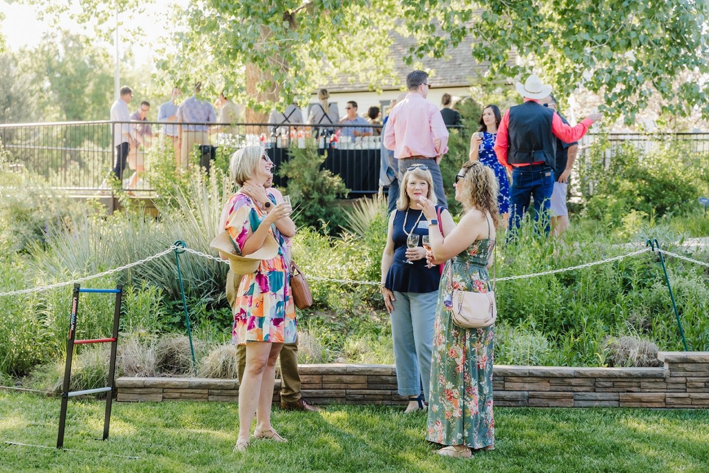 People socializing outdoors in a garden, some with drinks, near a low wall and a bridge.