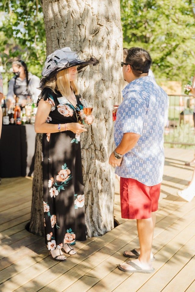 Couple socializing by a tree on a wooden deck. Woman in floral dress and hat; man in patterned shirt and red shorts.