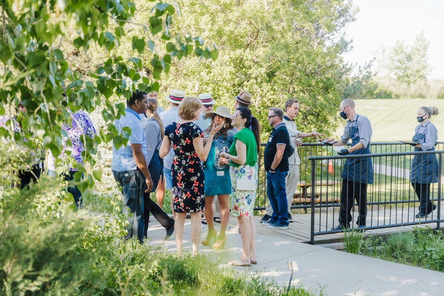 Group of people on a bridge, some conversing, near a grassy area. Two servers stand nearby.