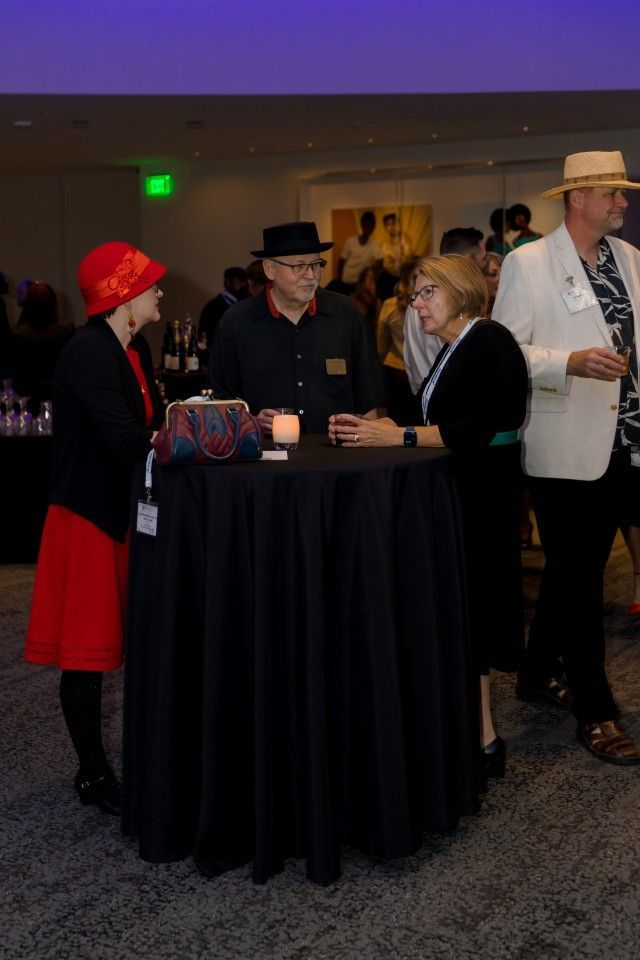 People conversing around a black cocktail table at an event. Some are wearing hats and formal attire.