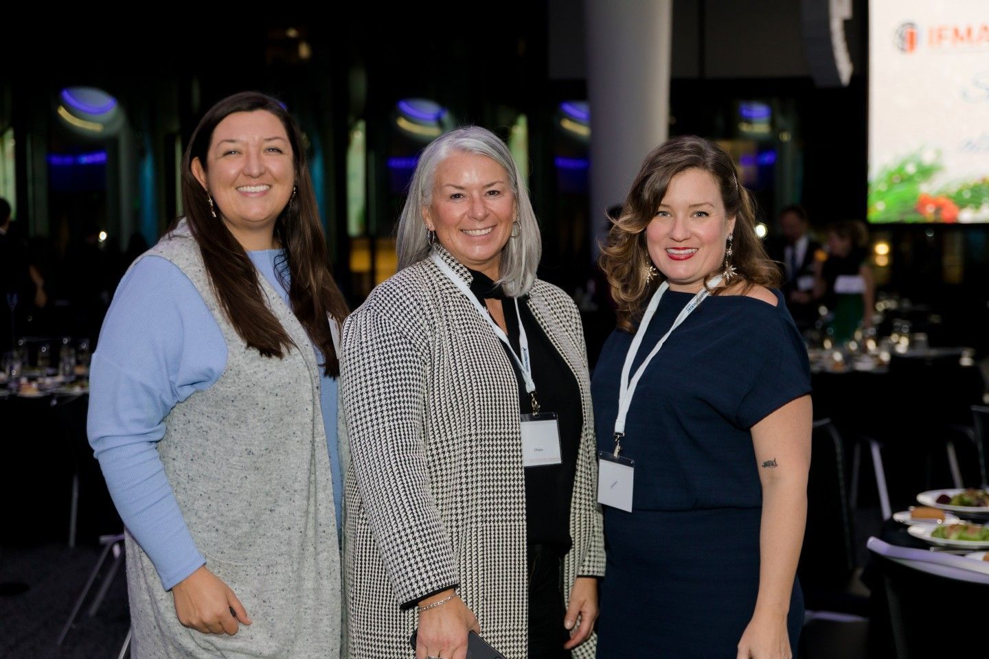 Three women smile at a gala event. Middle woman in patterned coat, others in blue, dark blue. Tables in background.