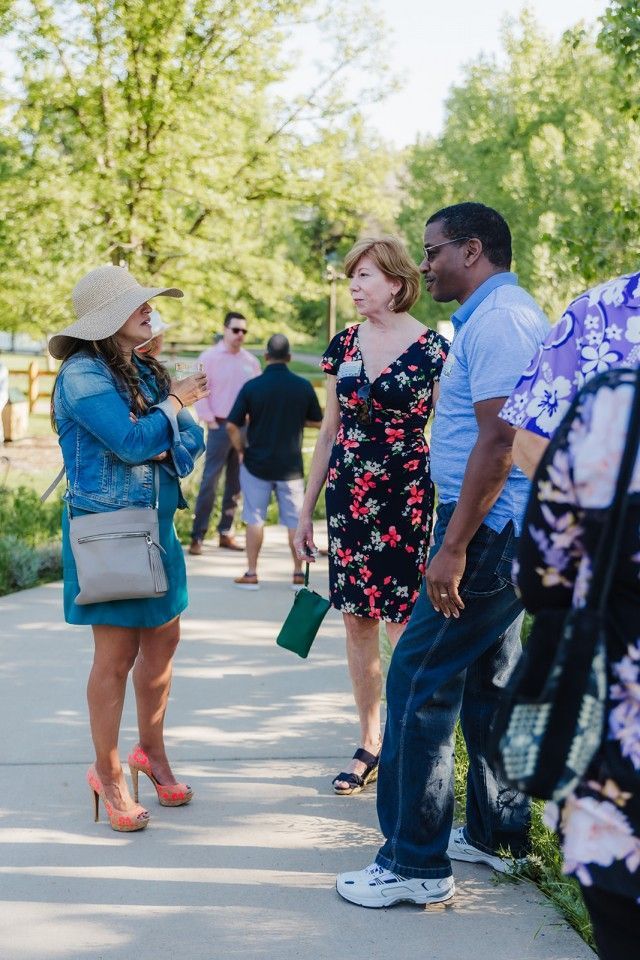 Group of people in casual attire outdoors, conversing. One woman wears denim jacket, hat, and heels. Others are nearby.