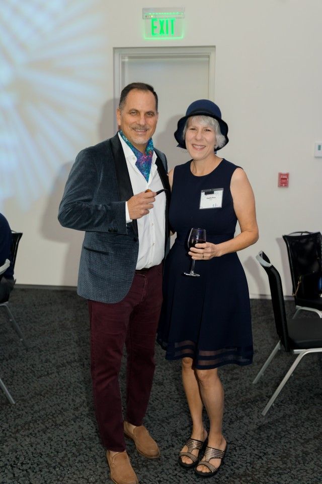 Man in velvet jacket and woman in a dress pose together; woman holds wine glass.