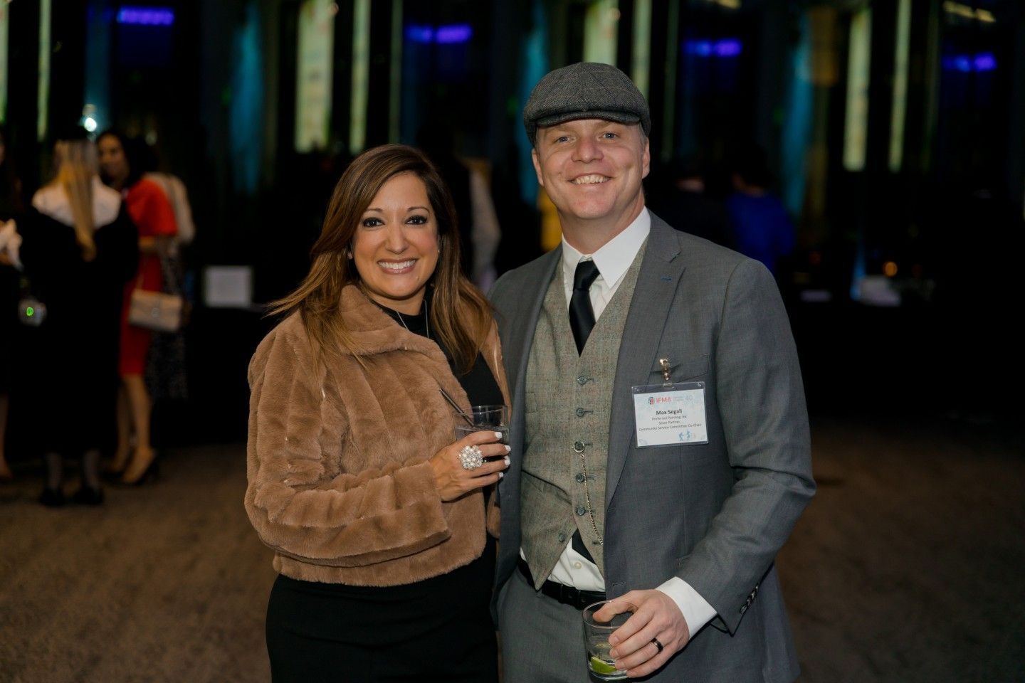 Woman in fur coat and man in tweed suit, smiling, holding drinks at an event.