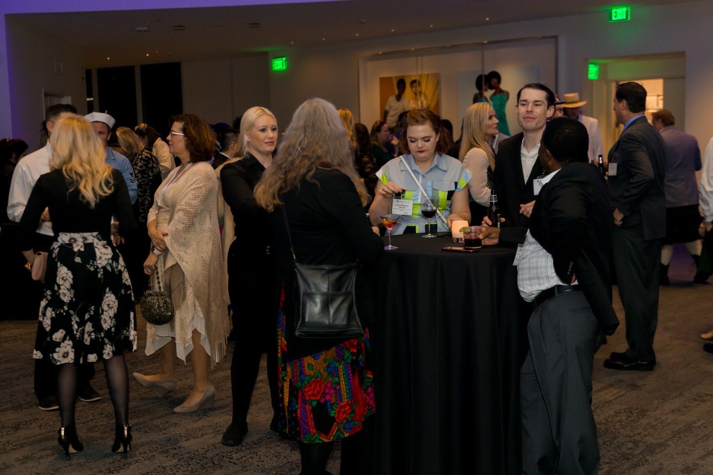 People at an event mingle, drink beverages, and converse at cocktail tables in a brightly lit room.