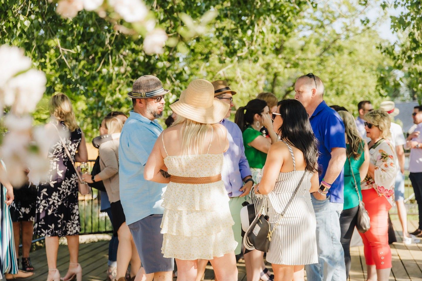 Guests mingling outdoors; woman in hat, light dress, brown belt; other people chatting, trees in background.
