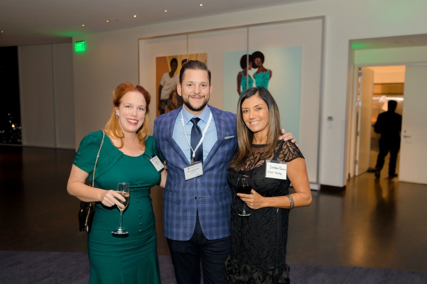 Three people at an event, holding drinks. Man in blazer, women in dresses pose. Modern building interior.