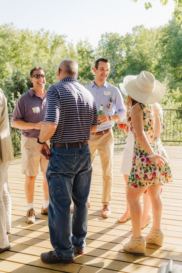 People socializing on an outdoor wooden deck with trees in the background. Some are holding drinks and smiling.