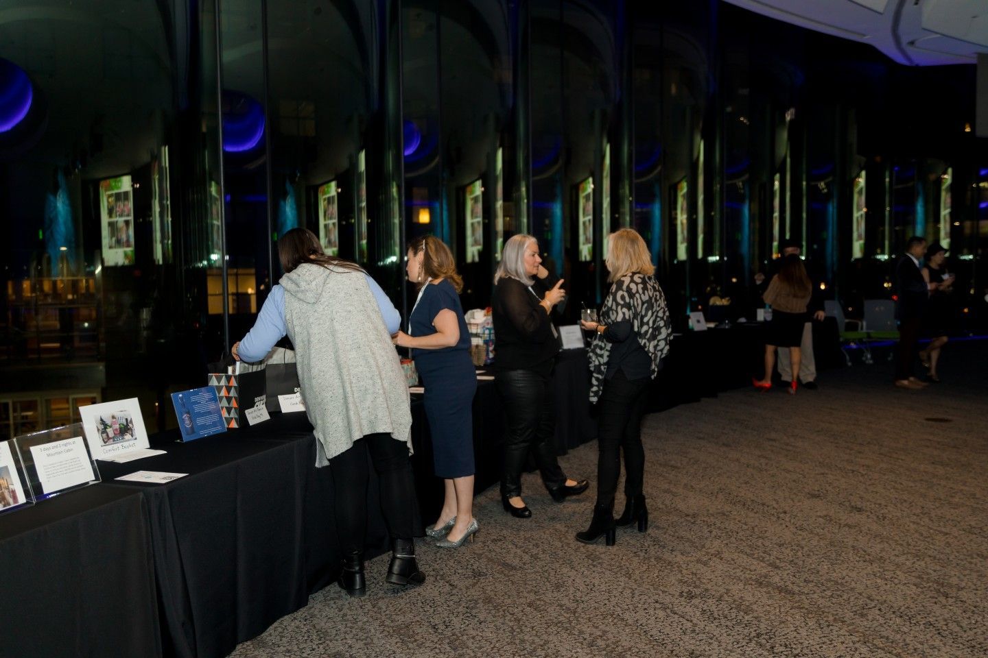 People at a formal event stand near a table with items, under a blue-lit ceiling.