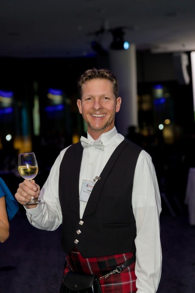 Man in formal wear with a kilt, holding a glass of white wine, smiling at an event.