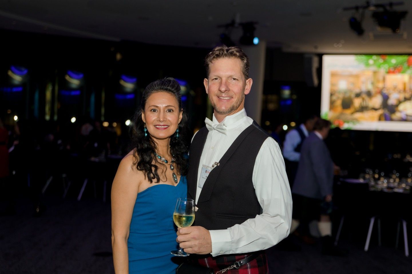 Woman in blue gown and man in formal wear, holding drinks, smiling at event.