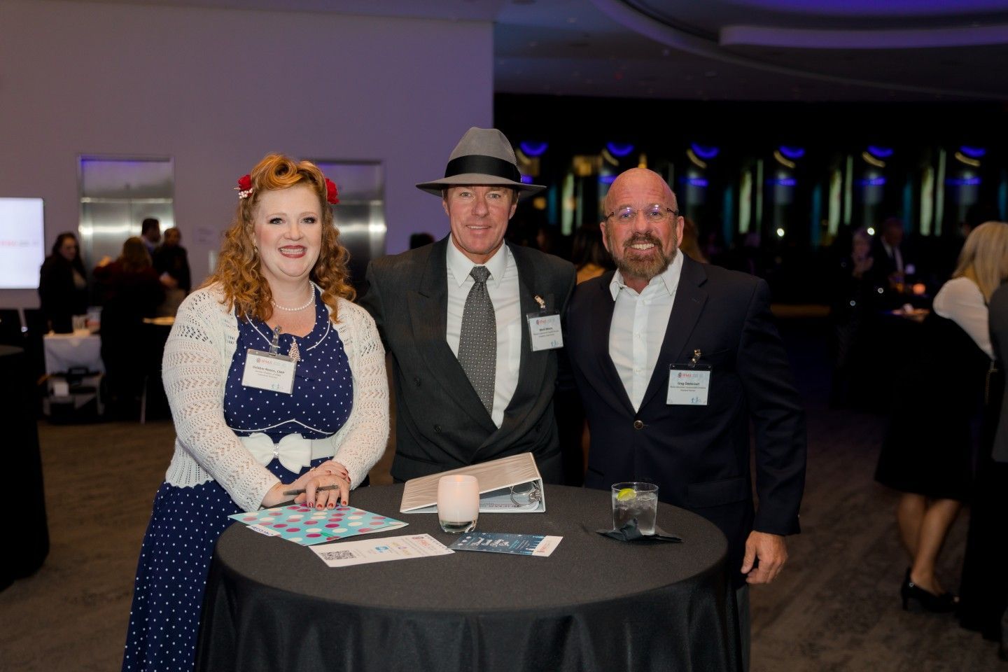 Three people at an event, posing near a table. One woman in polka dots, two men in suits.