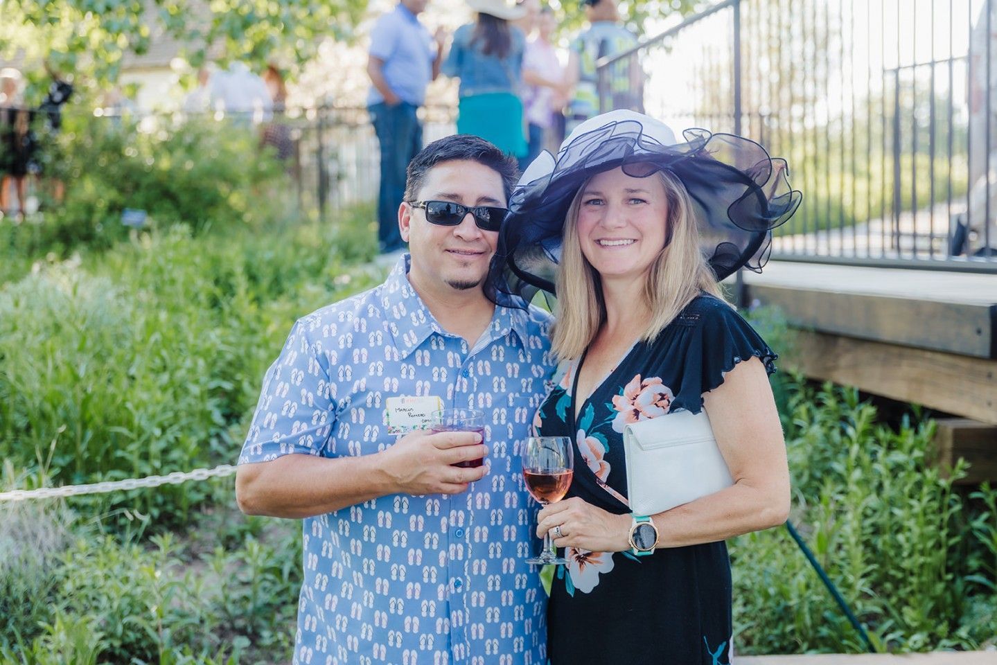 Man and woman smiling, holding drinks, outdoors. Woman wears a large hat, floral dress. Man in patterned shirt, sunglasses.