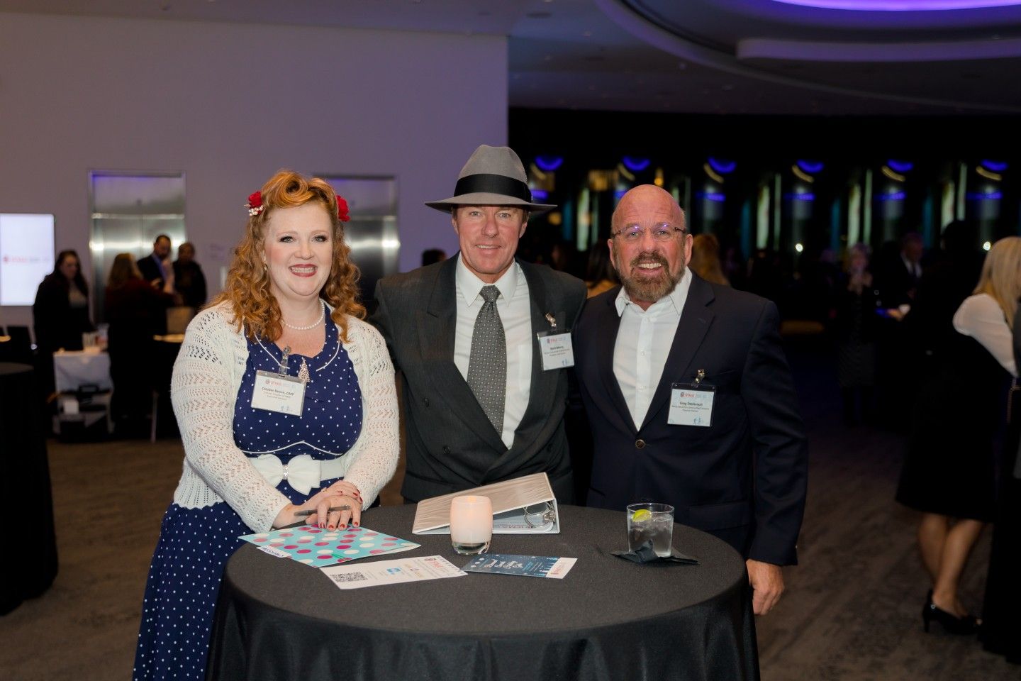 Three people smiling at a networking event, posing around a small table; woman in polka dots, man in fedora.