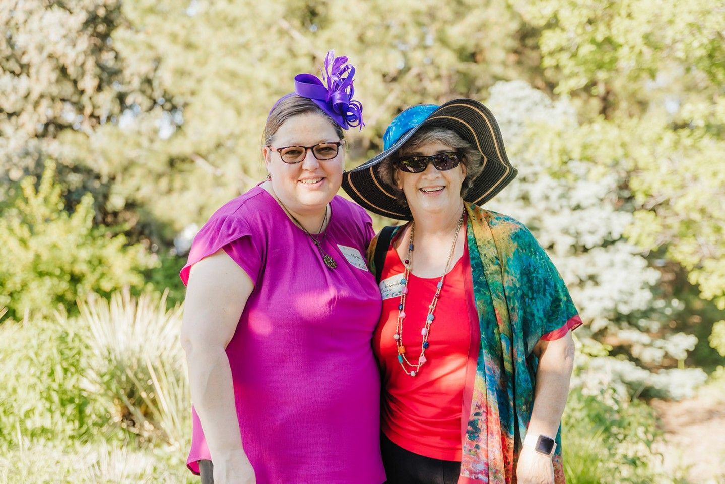 Two women smiling outdoors. One in a purple top and hat, the other in a red top and wide-brimmed hat.