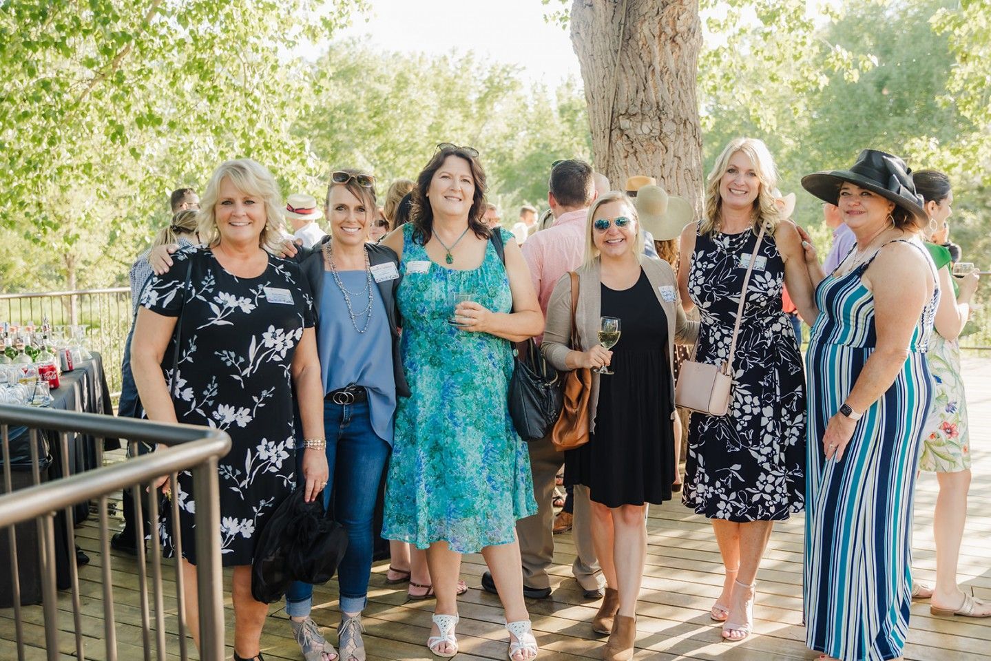A group of women smiling, posing for a photo outdoors. Some are holding drinks, a few are wearing nametags.