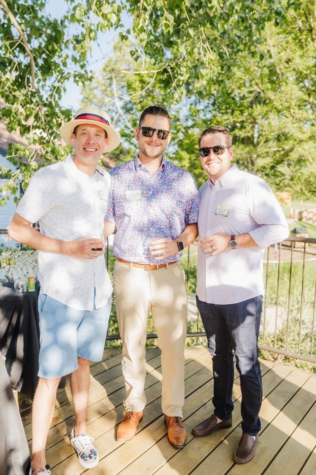 Three men standing on a wooden deck, smiling. Two wear sunglasses. Summer clothing.