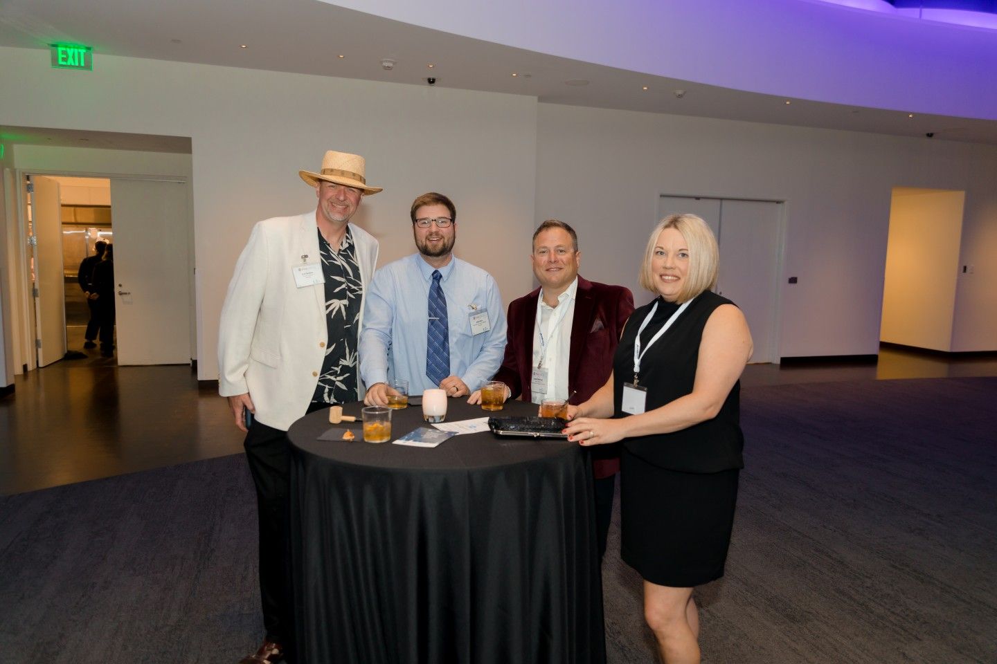 Four people standing around a small black table, drinks visible. The setting is an indoor event space with white walls.